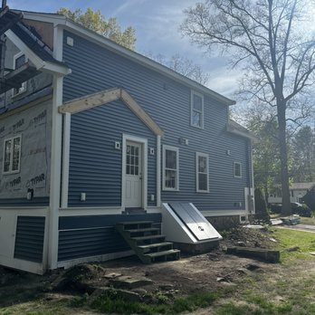 Gannon Construction - The image shows a two-story house with blue vinyl siding, multiple windows, and a small staircase leading to a white door. Part of the house is still under construction, with visible building materials and an unfinished section. The yard has dirt and grass, with a large tree and other houses in the background. The sky is clear with some clouds, and sunlight illuminates the scene. - General Contractors Near Me - Scituate, Massachusetts