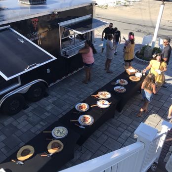 Shore Fire Pizza - Shore Fire Pizza (was Wicked Pizza) doing a private party in Sagamore Beach, MA. - A group of people is gathered outside a black food truck with a serving window, where a woman is placing an order. Several pizzas are displayed on black tablecloths on a table nearby. The scene takes place on a paved outdoor area with a white fence and residential buildings in the background, indicating an outdoor event or gathering. - Food Trucks Near Me