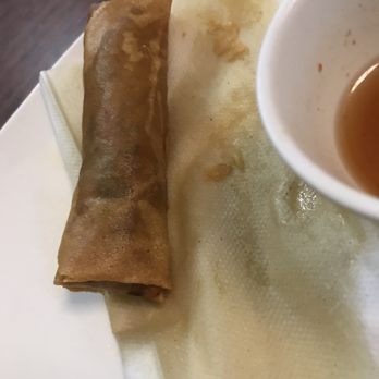Pho Saigon - A fried spring roll is placed on a white plate lined with a paper towel, with some crumbs and oil residue visible. A small cup containing a brown dipping sauce is partially visible in the upper right corner. The setting appears to be a restaurant or dining area, with a dark wooden table surface in the background. - Vietnamese Near Me - Tucson, Arizona
