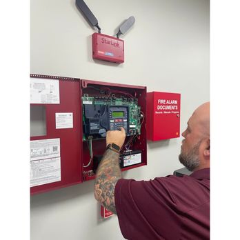 Spartan Fire Protection - Man with tattooed arm, wearing a maroon shirt, operates a fire alarm panel mounted on a wall, next to a red "Fire Alarm Docum... - Fire Protection Services Near Me - Houston, Texas Spartan Fire Protection - Man with tattooed arm, wearing a maroon shirt, operates a fire alarm panel mounted on a wall, next to a red "Fire Alarm Docum... - Fire Protection Services Near Me - Houston, Texas
