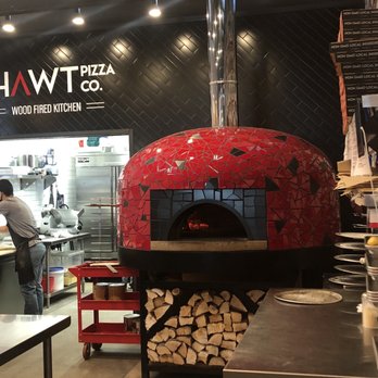 Hawt Pizza Co. - Where the magic happens - A red mosaic tile wood-fired pizza oven with a chimney pipe extending upward is positioned in a restaurant kitchen. The oven has a black opening with a visible fire inside. Below the oven, there is a storage area filled with chopped firewood. To the left, a person is working in the kitchen, and to the right, there are metal countertops and shelves with plates and utensils. The background features a black wall with the restaurant's logo and a glimpse into the kitchen area. - Pizza Near Me - Albuquerque, New Mexico