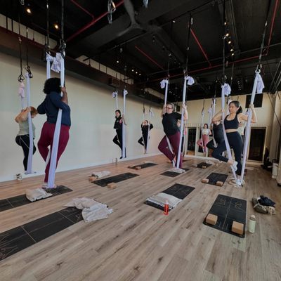 Aerial Yoga - Aerial yoga is a combination of yoga postures, pilates, gymnastics, and dance. With the use of a suspended silk hammock, which can also be thought of as a "yoga swing", this open level class will teach you to flow through yoga stretches with grace and offer the opportunity to practice poses that may not be as easily accessible on the mat.
Doors open 15 minutes before class starts.
Students should arrive no less than 10 minutes prior to class start, to leave time for hammock adjustment and check-in. Aerial Yoga - Aerial yoga is a combination of yoga postures, pilates, gymnastics, and dance. With the use of a suspended silk hammock, which can also be thought of as a "yoga swing", this open level class will teach you to flow through yoga stretches with grace and offer the opportunity to practice poses that may not be as easily accessible on the mat.
Doors open 15 minutes before class starts.
Students should arrive no less than 10 minutes prior to class start, to leave time for hammock adjustment and check-in.