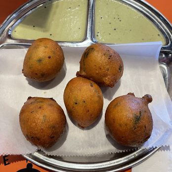 Malgudi Cafe - Goli bonda - Five fried Indian snacks, likely vada, are arranged on a paper-lined tray with green chutney in a three-compartment stainless steel dish. - Indian Near Me - Exton, Pennsylvania