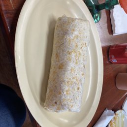 Burrito Pastor (Marinated pork) - A burrito wrapped in paper is placed on a beige oval plate at a restaurant table, with a glass of drink and other tableware nearby.