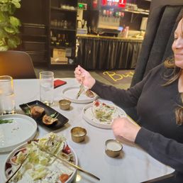 A woman sits at a table with plates of salad, glasses of water, and small bowls, in a restaurant with a bar area in the background.