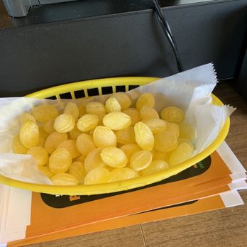 The Lemon Drop - Lemon drops at checkout - A yellow plastic basket filled with lemon drops candies, lined with white paper, is positioned on a table. The candies are round, yellow, and appear to be coated with a sugar-like substance. The basket is placed on top of a menu or paper placemat, and in the background, there is a black electronic device with cables connected to it, situated on a wooden surface. - Burgers Near Me - Anderson, Indiana