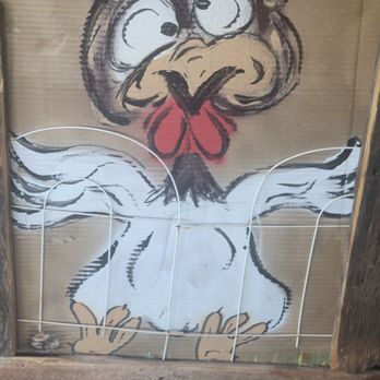 Rooster Run Cafe - A cartoon-style chicken is painted on a cardboard surface, with large, expressive eyes, a red comb, and a beak. The chicken's body is white with black outlines, and it has yellow feet. A white wire frame is placed over the painting, possibly for decorative or structural purposes. The background includes a wooden frame and a tiled floor. - Steakhouses Near Me - Hurricane, Utah