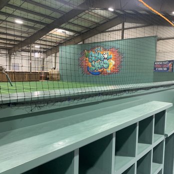 School Yahd - The image shows an indoor sports facility with a green court, a net, and overhead lighting. The ceiling is high with metal beams and a roof structure. A graffiti mural is visible on the wall, featuring colorful lettering. There is a green storage unit with cubbyholes in the foreground. The space appears to be used for recreational sports activities such as volleyball or badminton. - Recreation Centers Near Me - West Boylston, Massachusetts