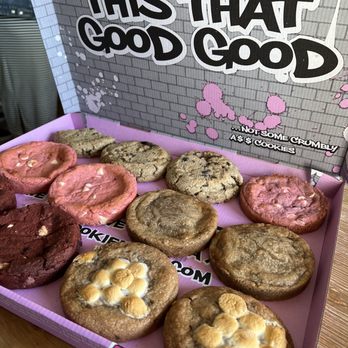 Cookie Plug - Firecracker Crunch Nug Cookie Cream Pink Elephant - The image shows a pink box filled with various cookies, including pink frosted cookies with white chips, chocolate cookies, and oatmeal cookies with white chocolate chips. The box is placed on a wooden surface, and the background features a decorative sign with graffiti-style text and pink paint splatters, indicating a bakery or cookie shop setting. - Bakeries Near Me - El Paso, Texas