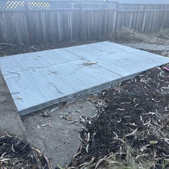 Method Remodeling - The large pad we had poured for our shed to sit on. - A concrete pad has been poured and set on a dirt surface in a backyard area, bordered by a wooden fence. The pad consists of multiple sections with visible joints, and there are scattered leaves and small debris around it. The surrounding area includes soil, some plants, and a garden hose on the right side of the image. - General Contractors Near Me - Burlingame, California