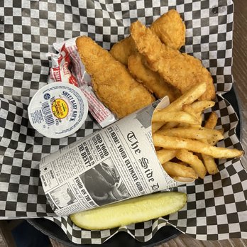 Coffee Bistro - Chicken strips and fries - Fried fish fillets, French fries, a pickle spear, and a small container of tartar sauce are arranged on checkered paper inside a basket. The fish fillets are golden brown and crispy, with the fries placed beside them. The pickle spear is at the front, and a small cup of tartar sauce is included for dipping. The basket is on a wooden table, suggesting a casual dining setting. - Coffee & Tea Near Me - Clovis, New Mexico