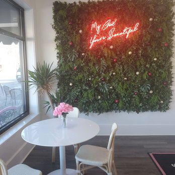 Shake It - Photo Op - A small seating area features a white round table with a pink flower arrangement, surrounded by white chairs, next to a window. - Juice Bars & Smoothies Near Me - Lexington, Kentucky