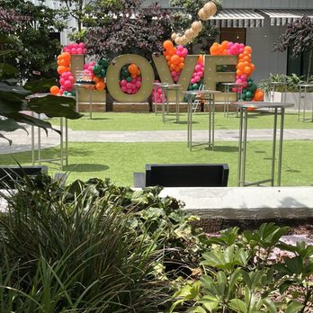 High/Low - Balloon arrangements in pink, orange, and green form the word 'LOVE' on a grassy outdoor area, decorated with hanging lanterns and surrounded by trees and plants. Several tall, round tables are positioned around the space, indicating a social gathering or celebration. The background features a building with large windows and striped awnings, creating a festive atmosphere for an outdoor event. - Breakfast & Brunch Near Me - Oceanside, California
