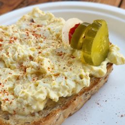 Egg Salad Sandwich - A slice of seeded bread with egg salad, garnished with pickles, served on a white plate at Southpine Bakery.