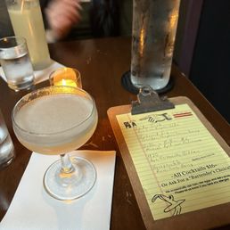 Fitzgerald - A cocktail in a coupe glass is placed on a white napkin on a wooden table. Next to it is a large, clear water bottle with condensation, and a menu on a clipboard with handwritten drink options. In the background, a person with long dark hair and a black top is partially visible, along with a glass of water and a lemon-infused drink with a garnish. The setting appears to be a bar or restaurant with dim lighting.