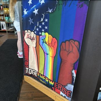 Java Central Cafe and Roaster - The banner features three raised fists in various skin tones and rainbow colors, symbolizing unity and diversity. The background includes a stylized American flag with stars and stripes, as well as a rainbow-colored backdrop representing LGBTQ+ pride. The phrase "TOGETHER WE RISE" is prominently displayed at the bottom in bold, multicolored letters. The banner is hanging on a counter in an indoor setting, with shelves and people visible in the background. - Coffee Roasteries Near Me - Westerville, Ohio
