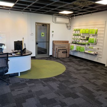 Extra Space Storage - Front office - The photo depicts the interior of a storage business office, featuring a reception desk with a computer and office chair on the left. The wall on the right has storage supplies, including boxes and rolls of packing material, displayed on a slatwall. The room has a carpeted floor with a green circular rug, a door at the back, and an air conditioning unit mounted on the wall. The ceiling has an open grid design with lighting fixtures. - Self Storage Near Me - Chicago, Illinois