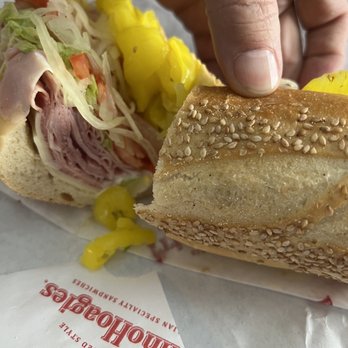 PrimoHoagies - American hoagie. - A person holds a sesame seed hoagie sandwich with visible layers of deli meats, lettuce, tomato, and pickles. The sandwich is partially wrapped in paper from PrimoHoagies, a sandwich shop in Bethlehem, PA. The image focuses on the sandwich and the hand holding it, with the background showing part of the wrapper and some of the sandwich ingredients. - Sandwiches Near Me - Bethlehem, Pennsylvania