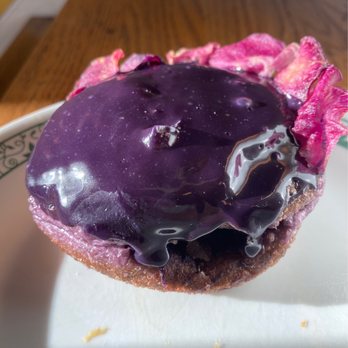 Kora - Ube donut - A pastry with a golden-brown crust is topped with a shiny, dark purple glaze that appears smooth and slightly reflective. Pink flower petals are arranged around the edges of the pastry, adding a decorative touch. The pastry is positioned on a white plate, with a wooden surface visible in the background. - Filipino Near Me - Sunnyside, New York