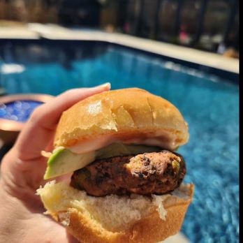 Cowlick - a hand holding a hamburger in front of a swimming pool - Food Trucks Near Me