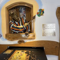 Hundertwasser Village - Friedensreich Hundertwasser leaning out of a wooden window and painting a colorful abstract mural on the wall