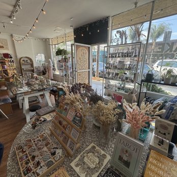 MAKE Collectives - The image shows the interior of a lifestyle shop with various shelves and tables holding jewelry, decorative items, and plants. Large windows at the front provide natural light, and there are string lights hanging from the ceiling. The store has a cozy, organized layout with a mix of modern and vintage decor, including potted plants and display stands. - Vinyl Records Near Me - Long Beach, California