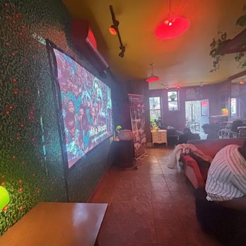 Red Rover - The image depicts a lounge with a cozy atmosphere, featuring velvet couches and a large screen on a textured wall displaying a group photo. The space has warm, dim lighting with red ceiling lights and some natural light coming through windows at the back. Several people are seated on couches, some engaged with their devices, creating a relaxed environment. - Coffee & Tea Near Me - Brooklyn, New York