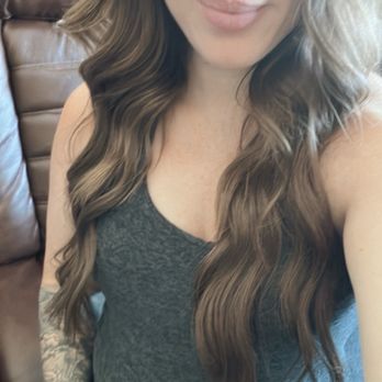 Arrow & Mane - 22in bellami extensions 1 row that came blonde but we died to my natural hair color after 1.5 years of use. - A woman with long, wavy brown hair and blue eyes is taking a selfie indoors, sitting on a brown leather couch, wearing a dark gray sleeveless top. - Hair Extensions Near Me - Tracy, California