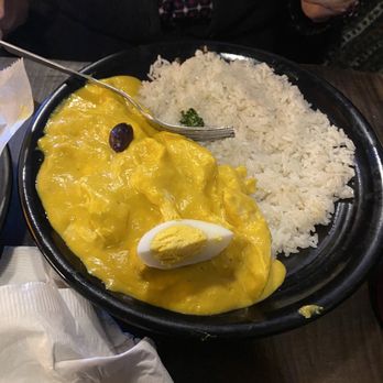 Culantro - Ají de Gallina - A black round plate holds a serving of yellow chicken curry with a halved boiled egg on top and a side of white rice. The curry appears creamy with a single dark olive or similar garnish. A fork rests on the plate, and a person in the background is holding a piece of bread or pastry. The setting suggests a casual dining environment. - Peruvian Near Me - Ann Arbor, Michigan