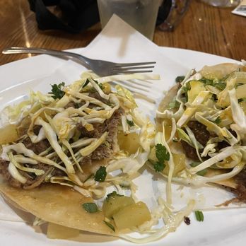 Quackers Lounge - Island Tacos - Two tacos are served on a white plate, topped with shredded cabbage, cilantro, and diced pineapple. The tacos contain a meat filling and are placed on a paper-lined tray. A fork rests on the plate, and a glass of water with ice is positioned behind the plate. The setting appears to be a casual dining environment, possibly a restaurant or cafe. - Sports Bars Near Me - Chico, California