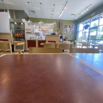 Uncle Henry - The image depicts the interior of Uncle Henry, a restaurant in Denver, CO, with several empty wooden tables and chairs arranged throughout the dining area. The ceiling has track lighting and hanging pendant lights. The counter area features a glass display with roasted chickens and a menu board on the wall. Large windows on the right side allow natural light to illuminate the space, which has a casual, minimal decor with a brick accent wall. - Asian Fusion Near Me - Denver, Colorado