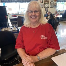 Barbar the owner! - A woman with blonde hair, glasses, and a red shirt is seated at a table in a restaurant, smiling at the camera.