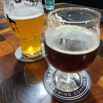 Ghost Hawk Brewing - Two glasses of beer, one light and one dark, are placed on a wooden bar table at a brewery, with a bar counter and glassware in the background. - Breweries Near Me - Clifton, New Jersey
