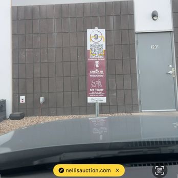 Nellis Auction - A sign featuring a QR code and text is placed on a stand outside a building with a gray wall and a door numbered 101. The sign provides instructions related to curbside pickup, check-in, and sitting tight, and is positioned near the ground with a small gravel area at the base. - Auction Houses Near Me - Mesa, Arizona