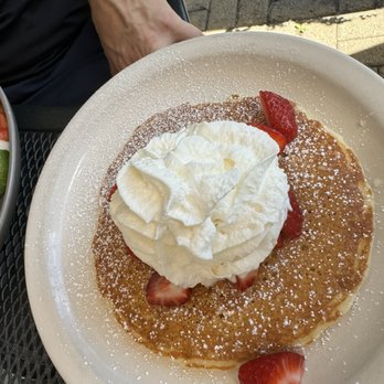 Breakfast House - SINGLE WW PANCAKE - A breakfast dish features a large pancake topped with a generous swirl of whipped cream and sliced strawberries. The pancake is dusted with powdered sugar and served on a white, round plate. Part of a salad with avocado and tomato is visible on the side, and the setting appears to be outdoors with a textured surface underneath. - Breakfast & Brunch Near Me - Chicago, Illinois