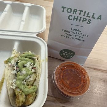 Tallboy Taco - A taco with toppings and sauce in a takeout container, accompanied by a bag of tortilla chips and a small cup of salsa on a wooden table. - Tacos Near Me - Chicago, Illinois