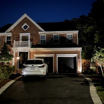 NOVA Landscape Construction - Repaved driveway with paver border - A two-story brick house at night with a driveway, illuminated by exterior lighting, featuring a white car parked outside and landscaped yard. - Snow Removal Near Me - Chantilly, Virginia