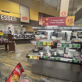 Kroger - Frozen food section at Kroger grocery store displaying pre-packaged meals and food items on refrigerated shelves. A sign advertises fresh chicken for $5, and other price tags are visible on different products. The store has a wooden floor and a yellow wall with a partial sign reading 'LICATESSEN.' The shopping cart in the foreground contains a bag of Italian seasoning. The area appears organized with clear pricing and product placement. - Grocery Near Me - Dearborn, Michigan