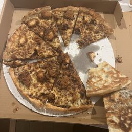 Yummmmmm - A pizza with cheese and meat toppings is placed in an open cardboard pizza box. Several slices have been taken, revealing the crust, cheese, and toppings. There are crumbs and small pieces of pizza on the box and nearby. The pizza appears to be a typical takeout or delivery style, with a golden-brown crust and melted cheese.