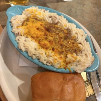 O'Niell's - Shepherds pie - A meal featuring shepherd's pie topped with melted cheese and mashed potatoes is presented on a blue oval dish. The dish is placed on a white tray, alongside a bread roll. In the background, there are glasses of water and a glass of beer on a wooden table, indicating a restaurant setting. - Irish Near Me - Albuquerque, New Mexico