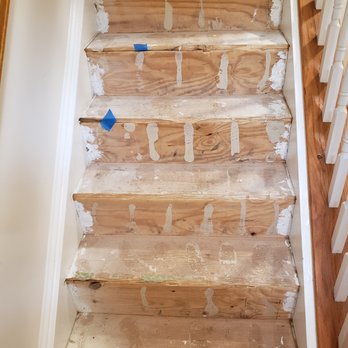 Carrillo Improvements - Before Photo, from the bottom of the staircase - The image shows a wooden staircase with several steps that are partially sanded or stripped, revealing the natural wood grain. Some steps have blue painter's tape, indicating areas for further work or protection. The sides of the staircase have white paint or primer, and the overall appearance suggests ongoing renovation or refinishing of the stairs. - Painters Near Me