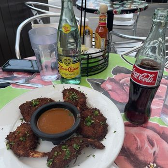Tune-Up Cafe - Coconut shrimp with spicy bbq sauce dip - A plate of fried shrimp garnished with herbs and served with a dipping sauce is placed on a table at an outdoor restaurant. The table also holds a Coca-Cola bottle, a bottle of mineral water, a glass of water, and condiments including ketchup and mustard. The setting includes metal chairs, a table with a floral tablecloth, and potted plants in the background, indicating an outdoor dining area. - New Mexican Cuisine Near Me - Santa Fe, New Mexico