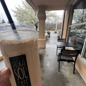 Mirasol's Cafe - Outside, large Chippi - A hand is holding a large iced coffee beverage from Mira Sol Café, with a black straw and a clear plastic lid. The drink appears creamy and light brown. The outdoor seating area of the café is visible, with two black tables and chairs, a sidewalk, and parked cars. The weather looks rainy, with wet pavement and overcast skies, and trees with budding leaves are in the background. - Coffee & Tea Near Me - Dartmouth, Massachusetts