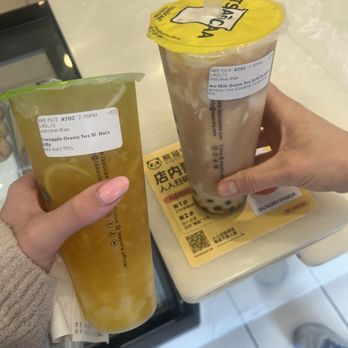Tsaocaa - Two bubble tea drinks are held by two individuals at a counter in a bubble tea shop. The drink on the left contains lemon slices and appears to be pineapple green tea, while the drink on the right has tapioca pearls and is labeled as green tea with milk. Both drinks are in clear plastic cups with sealed lids and straws. The background shows a white counter with a menu or promotional material and part of the shop interior. - Bubble Tea Near Me - Pittsburgh, Pennsylvania