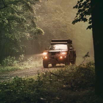 Nova Speed Shop - A vehicle with its headlights illuminated is driving along a dirt road through a dense forest. The scene is set at dusk or early evening, with soft, diffused light filtering through the trees. The vehicle appears to be an off-road or adventure vehicle, equipped with additional lights and gear on the roof. The surrounding environment includes tall trees, lush green foliage, and a misty or foggy atmosphere, creating a sense of wilderness and exploration. - Auto Customization Near Me - Manassas, Virginia