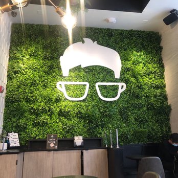 Gregorys Coffee - The image shows an interior cafe with a vibrant green wall made of plants, featuring a white logo of a hippo wearing glasses. The logo is illuminated and centered on the wall. There is a black ceiling shelf with decorative plants above the logo, and a modern light fixture with exposed bulbs hanging from the ceiling. The cafe has a counter with a sign indicating hiring, and seating includes a small round table and chairs. A person wearing a mask is seated on the right side of the image. - Coffee & Tea Near Me - Washington, DC, District of Columbia