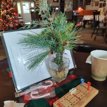 Bucky's - Centerpiece - The image shows a table in a cafe decorated for Christmas, with a glass vase holding holiday greenery including pine and cedar branches. A Tins game with two dice and colored pegs is on the table, along with candy canes in a plastic bag. A paper cup and a menu or sign are also visible. In the background, a Christmas tree with lights and ornaments, along with various wall decorations, create a festive atmosphere. - Breakfast & Brunch Near Me - Milton, New Hampshire