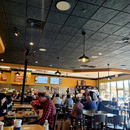 View from my table - The image shows the interior of a restaurant with multiple customers seated at tables, some engaged in eating and conversation. The ceiling is black with recessed lighting and hanging pendant lights. Large windows on the right side allow natural light to illuminate the space. In the background, a kitchen area is visible with staff working, and digital menu screens are mounted on the wall above the counter. The overall setting appears lively and well-lit.