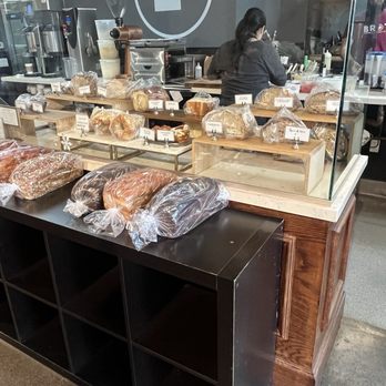 Brot Coffee - Homemade bread/mini bakery - Bakery display featuring several loaves of bread wrapped in plastic, placed on a black shelf and a wooden display stand. The counter has a glass barrier, and behind it, a barista is working with bakery equipment and supplies. The setting appears to be inside a bakery or coffee shop. - Coffee & Tea Near Me - Orange, California
