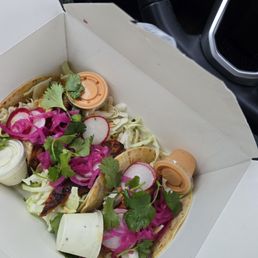 Two tacos with grilled meat, radish slices, pickled vegetables, cilantro, and sauces in a takeout container.